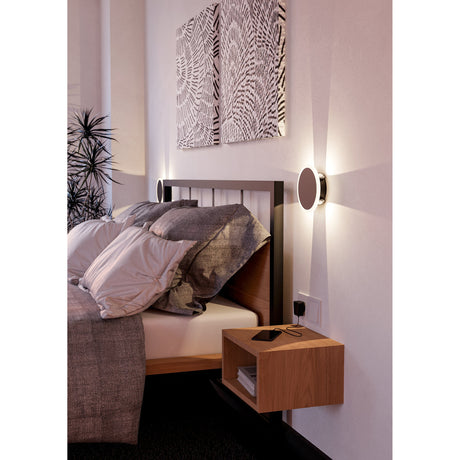 Talamello 1 LED Wall Light 17cm in black, round disc design with white diffuser ring, shown mounted beside a bed casting upward and downward light beams.