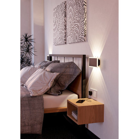 Talamello 1 LED Wall Light, 23cm black steel rectangular fixture with pale edge trim, mounted beside a bed casting warm upward and downward light in a bedroom setting.