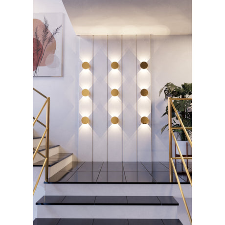 Talamello LED Wall Light in brushed gold, round 15cm aluminium discs arranged in rows on a staircase wall, casting warm up-and-down light beside a plant and framed artwork.