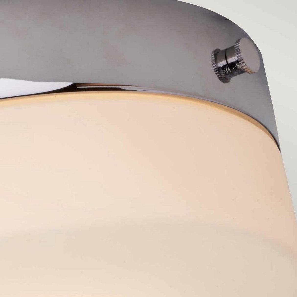 A close-up of the Tamar 1 Light Medium Flush in polished chrome, showcasing its modern design with an opal glass diffuser. The fixture includes a small screw detail on the metal component, while the soft LED lamp emits a warm glow.
