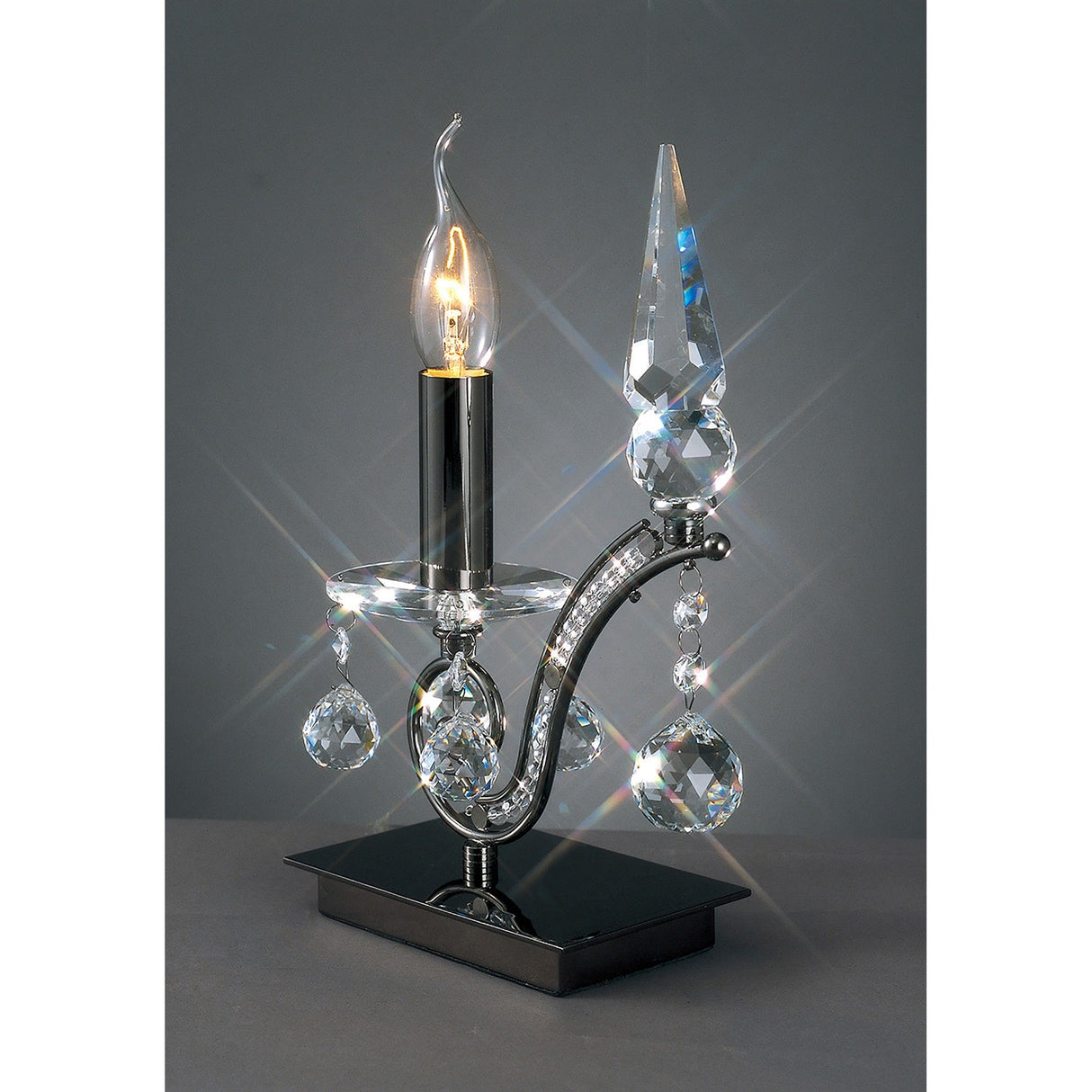 The Tara 1 Light Crystal Table Lamp - Black Chrome showcases a candle-shaped bulb, hanging crystal accents, and a pointed finial on a reflective base, ideal for adding elegant decorative lighting to your space.