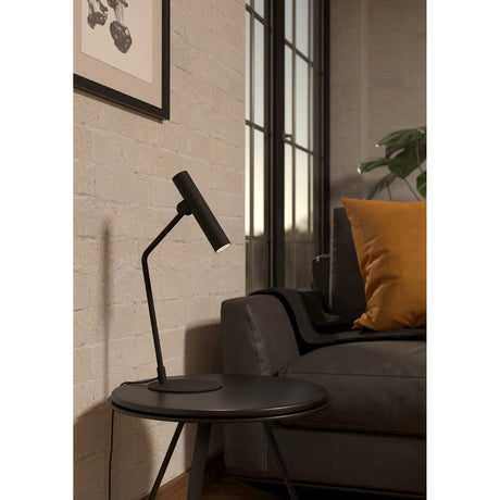 Tasmenne LED Table Lamp in black steel with a bent stem and pivoting cylindrical head, shown on a round side table beside a dark sofa and mustard cushion.