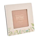 White ceramic square photo frame with 4x4in aperture, glossy finish, and wildflower sprigs in pink, yellow and blue along the bottom edge, on a plain background.