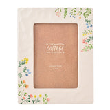 White ceramic photo frame with wildflower and leafy sprig details in pink, yellow and blue, holding a 4 x 6in photo, shown on a plain background.