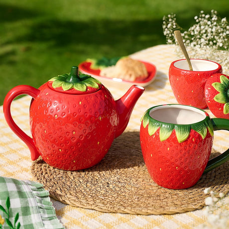 Strawberry Glazed Ceramic Teapot 820ml with Leaf Lid in Red, shaped like a berry with textured seed dimples, shown with matching strawberry mugs on an outdoor picnic table.