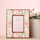 Rectangular wooden photo frame for a 6 x 4in photo, white face with pastel pink, peach and blue floral print and warm wood inner border, shown on a table beside flowers.