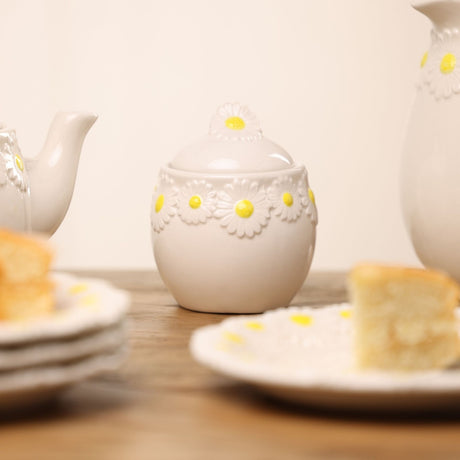 Round white stoneware sugar bowl with lid, featuring a raised daisy border and daisy-shaped knob with yellow centres, shown on a table with teapot and cake.