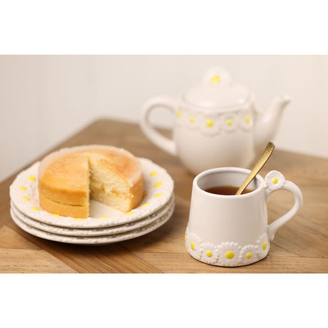 White stoneware Daisy Mug with raised daisy trim and flower-topped handle, shown with tea and a gold spoon alongside a matching teapot and stacked side plates with cake.