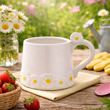 White stoneware mug with raised daisy details around the base and a daisy accent on the curved handle, shown outdoors on a wooden surface with strawberries and flowers.