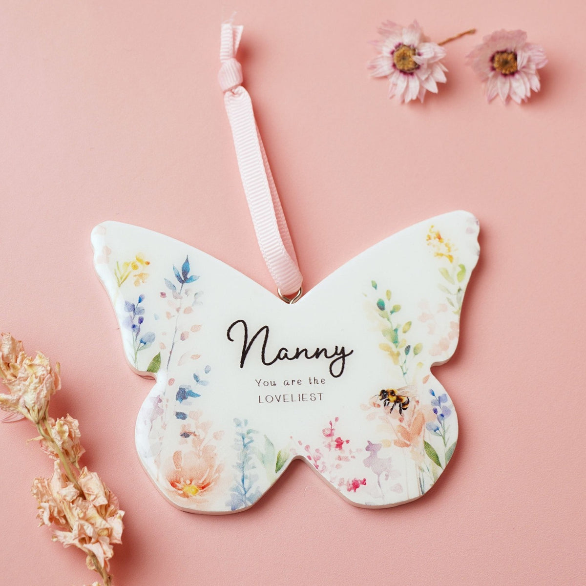 White wooden butterfly-shaped hanging decoration with pastel watercolour floral print, reading "Nanny, You are the LOVELIEST", on a pink ribbon against a pink background.