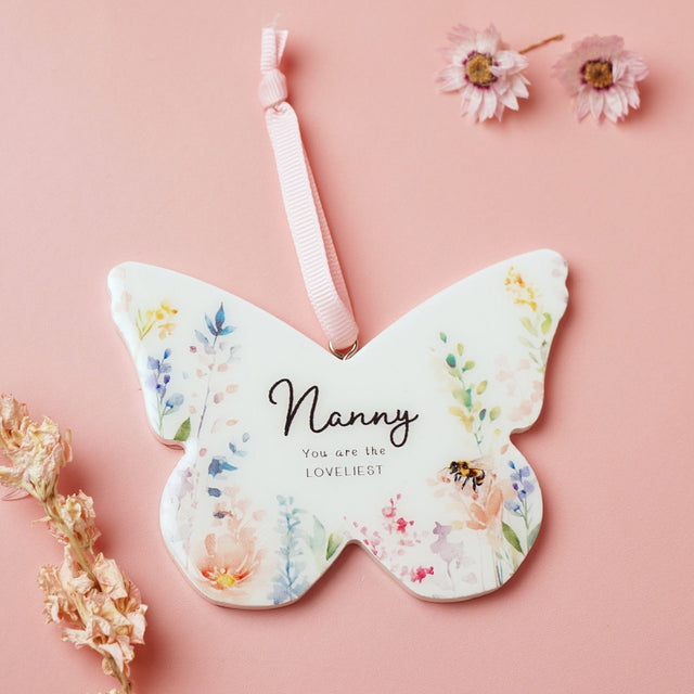 White wooden butterfly-shaped hanging decoration with pastel watercolour floral print, reading "Nanny, You are the LOVELIEST", on a pink ribbon against a pink background.