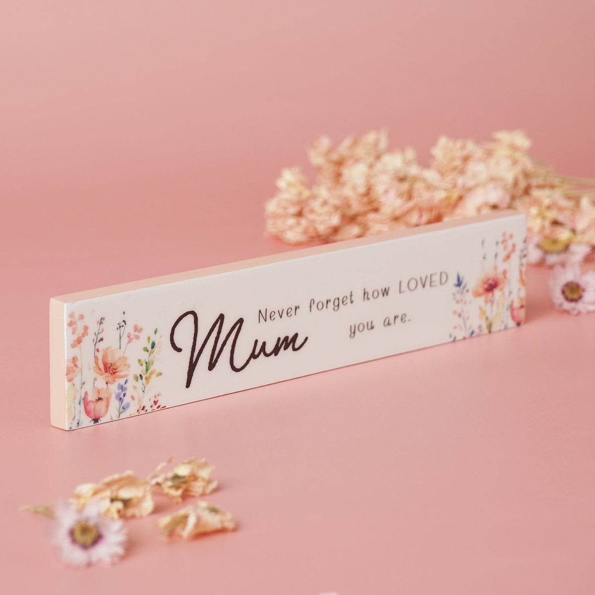 Slim white wooden plaque reading "Mum, Never forget how LOVED you are" in black script, flanked by watercolour pink and coral floral sprays, displayed on a pink background with dried flowers.