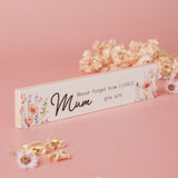 Slim white wooden plaque reading "Mum, Never forget how LOVED you are" in black script, flanked by watercolour pink and coral floral sprays, displayed on a pink background with dried flowers.