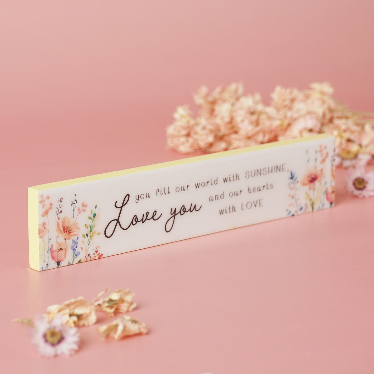 Slim wooden plaque with cream background and yellow edge, reading you fill our world with sunshine and our hearts with love in black script, surrounded by watercolour wildflowers on a pink surface.