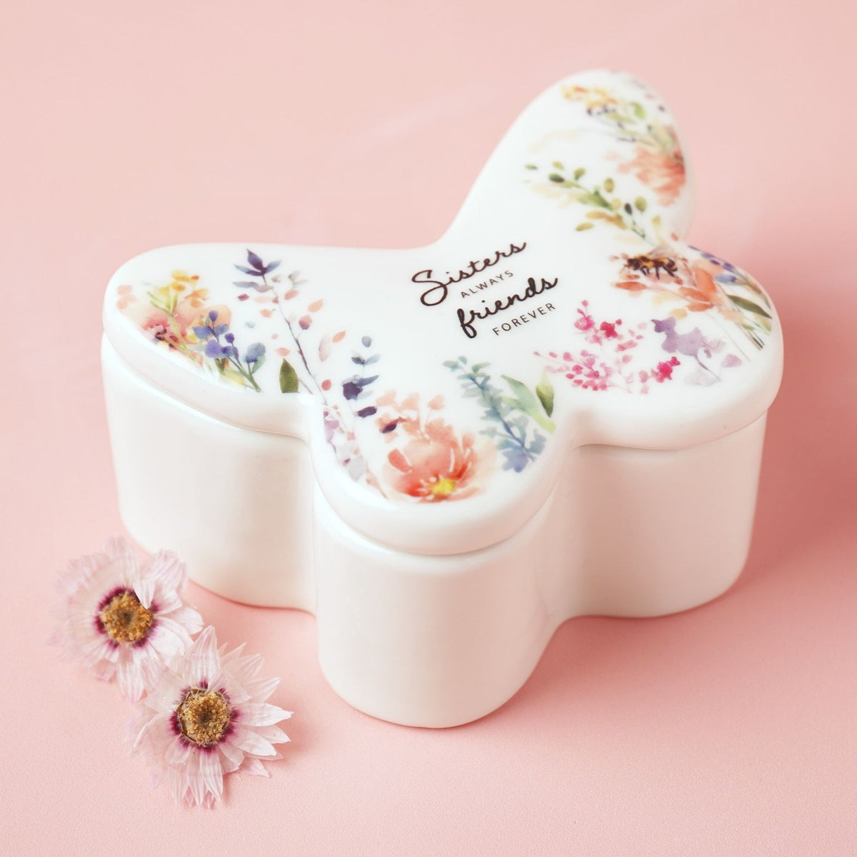 White glass butterfly-shaped trinket box with lid, featuring a watercolour floral print and "Sisters Always Friends Forever" script, shown on a pink background with dried flowers.
