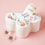 White glass butterfly-shaped trinket box with lid, featuring a watercolour floral print and "Sisters Always Friends Forever" script, shown on a pink background with dried flowers.