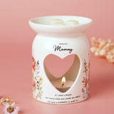 White glazed wax melt burner with heart cut-out, printed wildflowers and "Loveliest Mummy" text, lit tealight inside and wax melts on top, pink background.