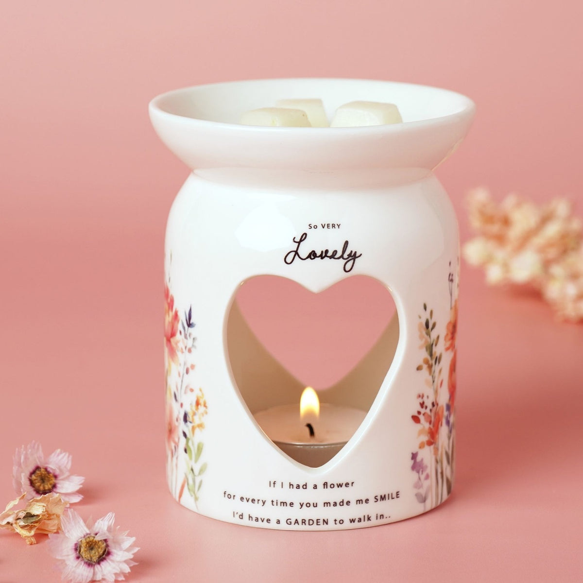 White glass wax melt burner with heart cut-out and lit tealight, printed "So Very Lovely" script and floral verse, wildflower design, on pink background.