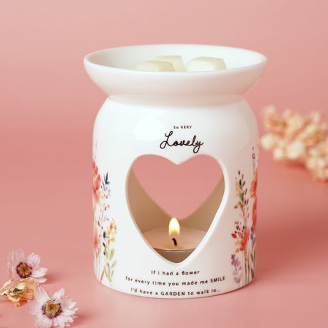 White glass wax melt burner with heart cut-out and lit tealight, printed "So Very Lovely" script and floral verse, wildflower design, on pink background.