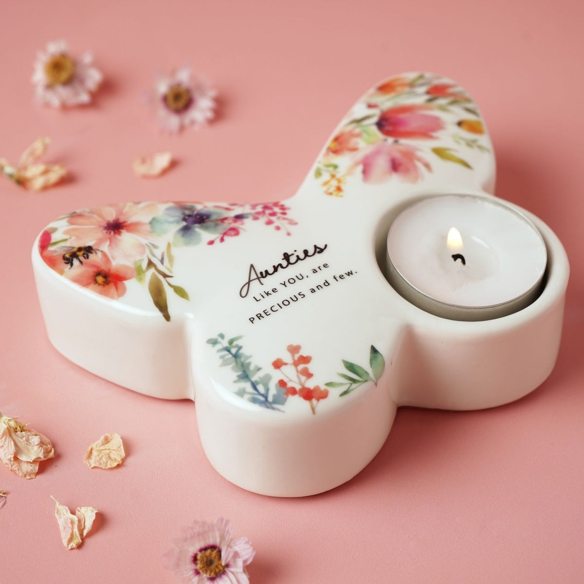 White glass butterfly tealight candle holder printed with watercolour florals and "Aunties Like YOU, are PRECIOUS and few", holding a lit tealight on a pink surface with scattered dried flowers.