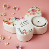 White glass butterfly tealight candle holder printed with watercolour florals and "Aunties Like YOU, are PRECIOUS and few", holding a lit tealight on a pink surface with scattered dried flowers.