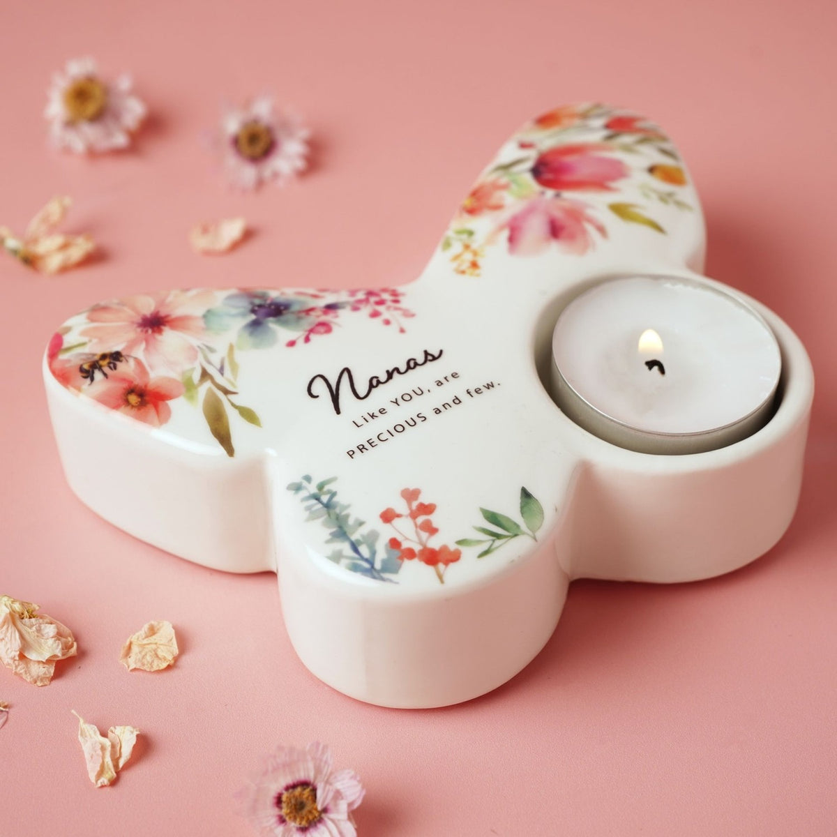 White glass butterfly-shaped tealight candle holder with painted floral design and "Nanas Like YOU, are PRECIOUS and few" text, lit tealight in wing cup, on pink background.