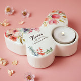 White glass butterfly-shaped tealight candle holder with painted floral design and "Nanas Like YOU, are PRECIOUS and few" text, lit tealight in wing cup, on pink background.