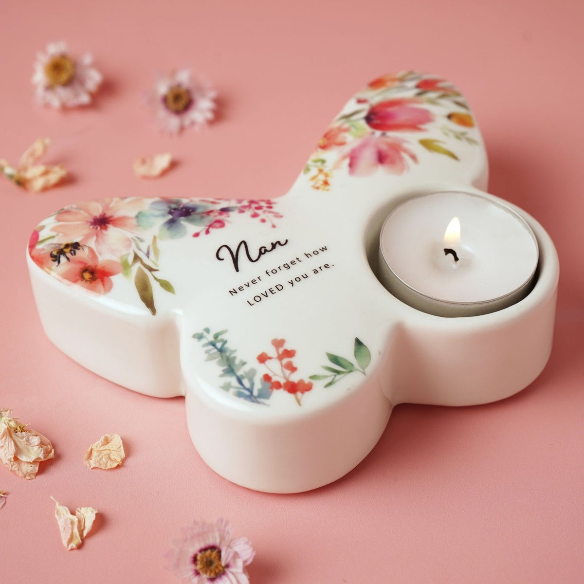 White glossy butterfly-shaped tealight candle holder with pink, coral and lilac watercolour florals, a bumblebee motif, and "Nan" text, lit tealight in one wing, on a pink background.