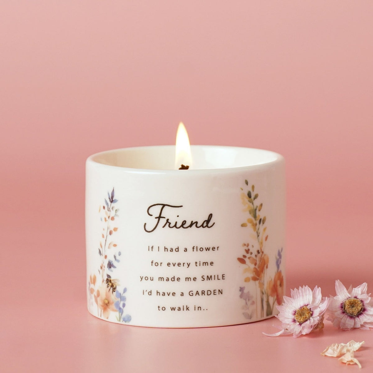 Round ceramic scented candle in off-white with wildflower print and "Friend" quote in script lettering, lit wick, shown against a pink background.