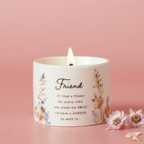Round ceramic scented candle in off-white with wildflower print and "Friend" quote in script lettering, lit wick, shown against a pink background.