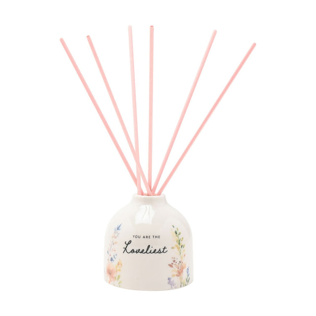 Off-white ceramic reed diffuser with rounded body, "You are the Loveliest" script and watercolour floral print, topped with six pink reeds, on a plain white background.