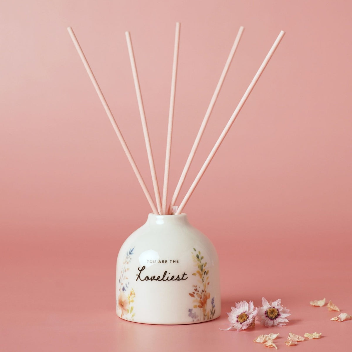 Off-white ceramic reed diffuser with rounded body, "You Are The Loveliest" script and watercolour wildflower print, five pink reeds, set against a pink background.