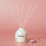 Off-white ceramic reed diffuser with rounded body, "You Are The Loveliest" script and watercolour wildflower print, five pink reeds, set against a pink background.
