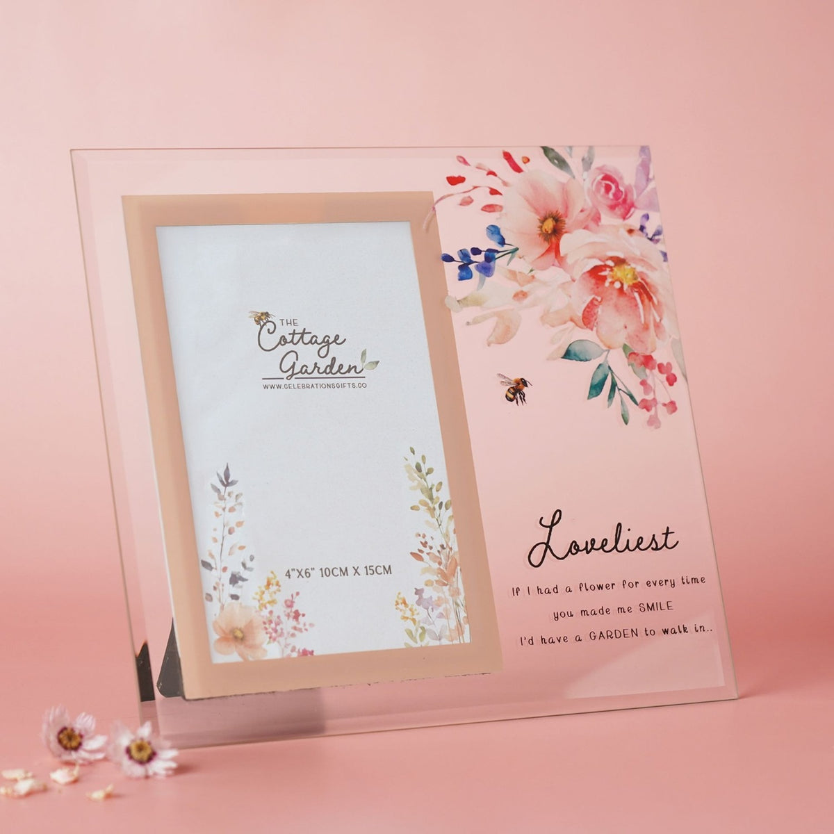 Freestanding glass photo frame with 4"x6" portrait opening, peach-pink border, watercolour floral and bee design, and "Loveliest" script text, shown against a pink background.