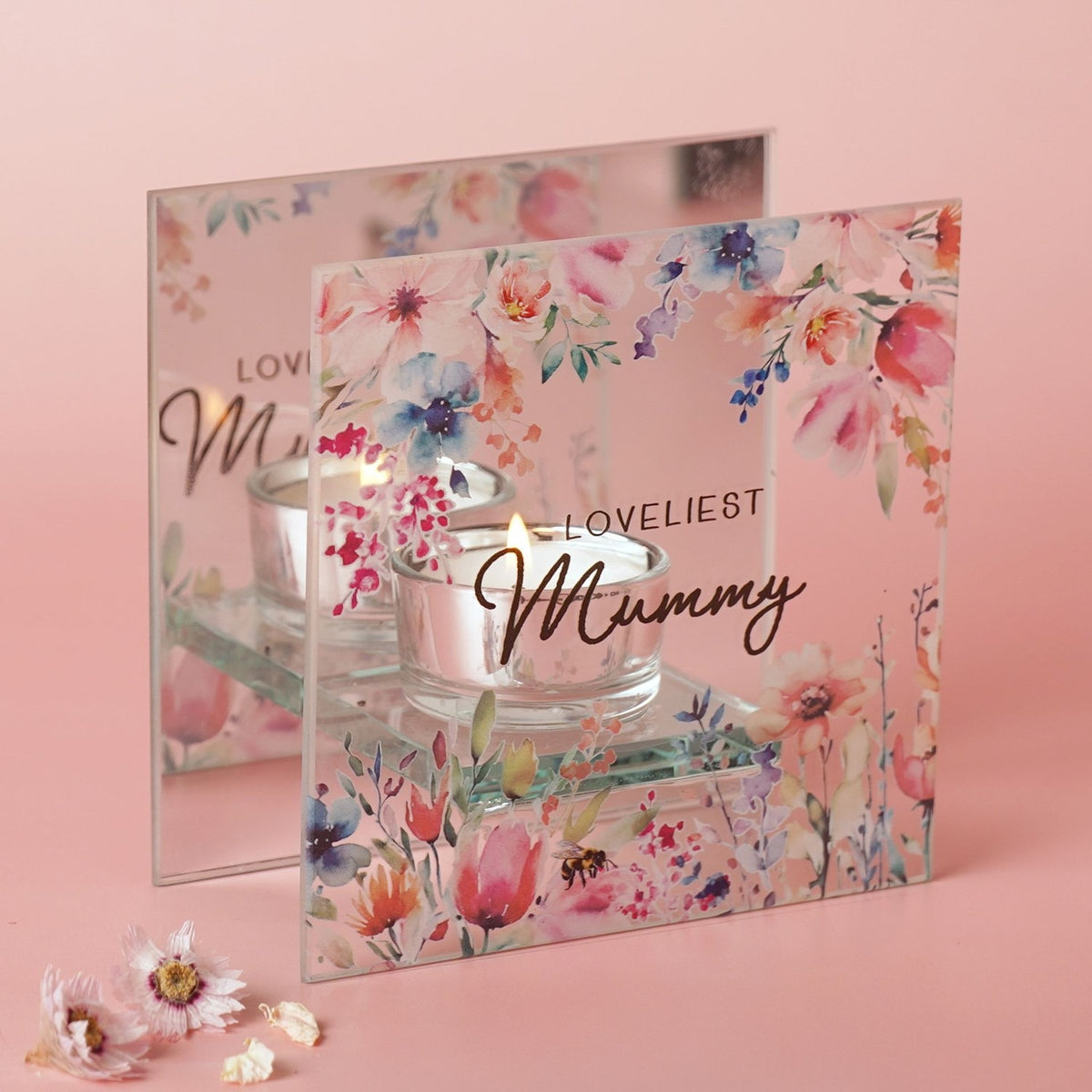 Square clear glass tealight candle holder printed with pastel floral watercolour blooms and "LOVELIEST Mummy" text, holding a lit candle on a glass base against a pink background.