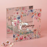 Square clear glass tealight candle holder printed with pastel floral watercolour blooms and "LOVELIEST Mummy" text, holding a lit candle on a glass base against a pink background.
