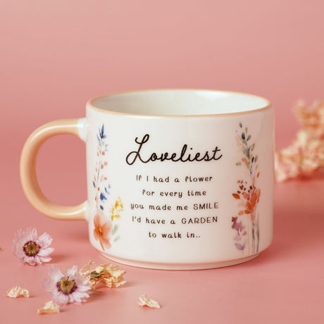 White stoneware mug with peach handle, printed with "Loveliest" script, a verse, and watercolour wildflowers, set against a pink background with dried flowers.