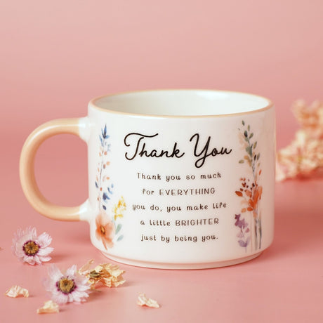 Off white stoneware mug with peach handle, printed with "Thank You" script and a thank you message, flanked by watercolour-style wildflowers, set against a pink background with dried flowers.