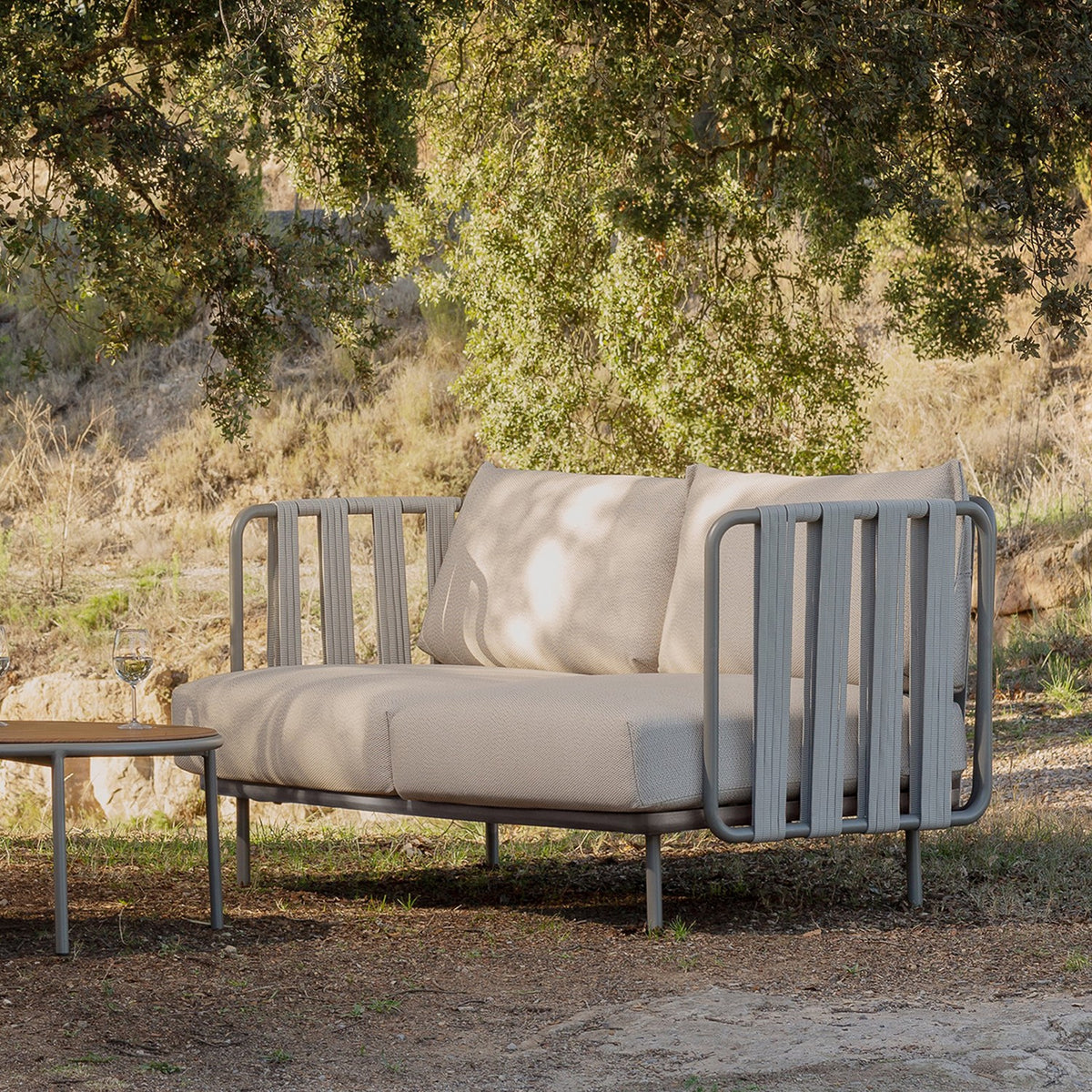 Teja Rope 2 Seater Garden Sofa with beige cushions and grey rope-wrapped aluminium arms, shown outdoors on a wooded garden terrace beside a matching side table.