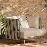 Teja Rope Garden Armchair with beige cushions, anthracite aluminium frame and woven rope side panels, shown outdoors against a stone wall with a palm plant.