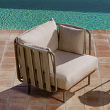 Teja Rope Garden Armchair with tubular aluminium frame wrapped in beige rope and cream cushions, set poolside on terracotta tiles beside turquoise water.