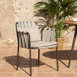 Teja rope garden dining armchair with black aluminium frame, woven rope sides and back, and beige seat and back cushions, shown on a patio beside a plant and table.