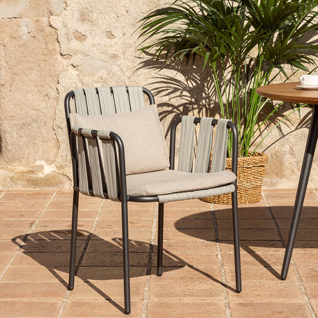 Teja rope garden dining armchair with black aluminium frame, woven rope sides and back, and beige seat and back cushions, shown on a patio beside a plant and table.