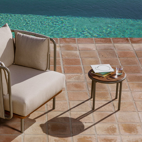 Teja round garden side table in aluminium with a wood-effect top and olive-green frame, set poolside beside a beige lounge chair with a book and glass on top.