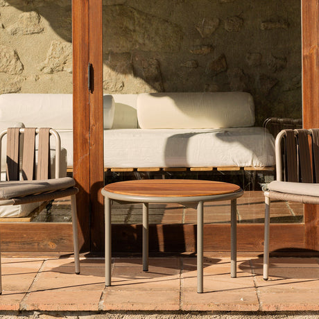 Teja Aluminium Round Garden Side Table, Ø60cm, with wood-effect top and beige round legs, shown on a terrace beside outdoor chairs and a sofa.