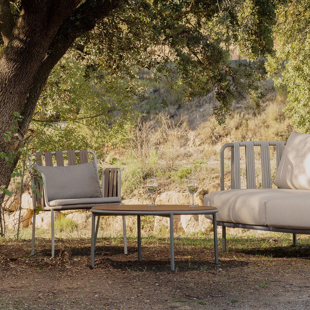 Teja round outdoor coffee table, Ø90cm, with wood-effect top and slim grey aluminium frame, set in a garden beside an armchair and sofa under tree shade.
