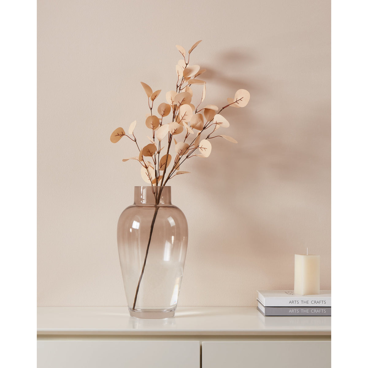 Tepaling artificial leaf branch in beige with round eucalyptus-style foliage on a thin brown stem, displayed in a glass vase on a sideboard against a pale pink wall.