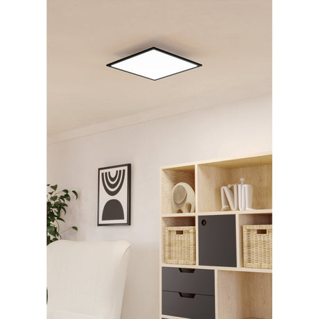 Tesvelle square LED ceiling panel light, 39.3 x 39.3cm, with slim black steel frame and white diffuser, shown recessed into a living room ceiling above shelving and a plant.