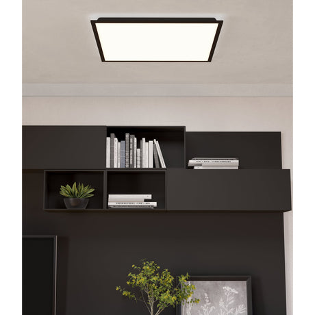 Tesvelle square LED ceiling panel light with slim black steel frame and white diffuser, shown installed in a living room above a dark wood shelving unit.
