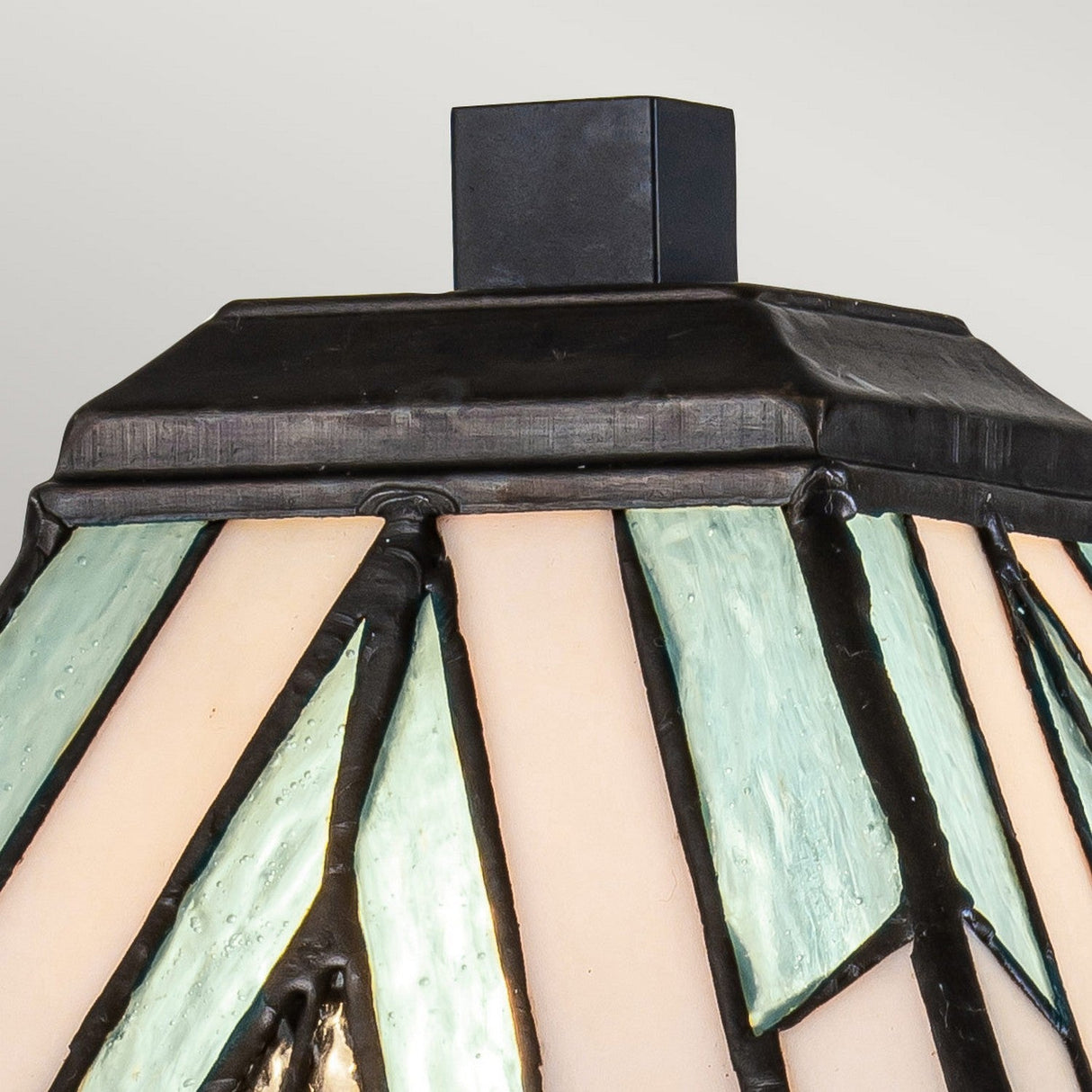 Close-up of the Alderley 1 Light Medium Table Lamp - Vintage Bronze, featuring a Tiffany-style lampshade with geometric designs in pale green and cream, framed by a black metal structure, against a plain gray background, evoking the essence of an art deco table lamp.